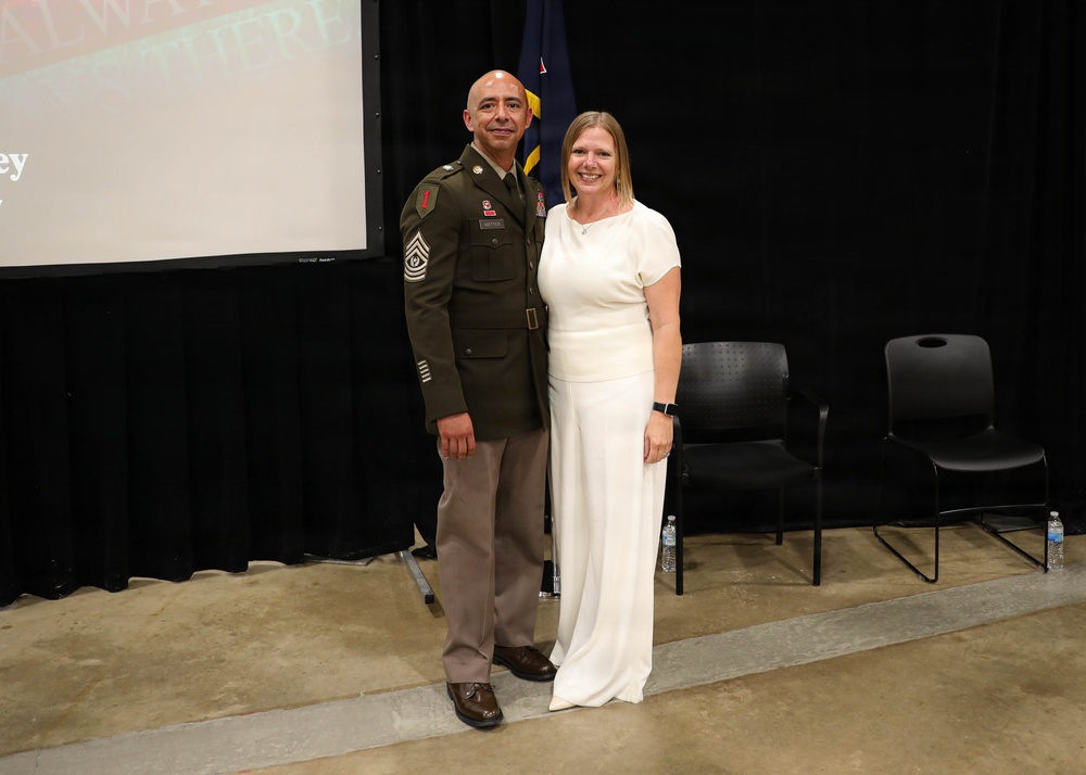 Two individuals pose together for a photo at what appears to be a formal military ceremony or event. One person is wearing a U.S. Army dress green uniform with visible rank insignia consistent with a Senior Non-Commissioned Officer, while the other is dressed in a white outfit. They are standing in front of a projection screen and a U.S. flag on a stage.