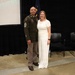 Two individuals pose together for a photo at what appears to be a formal military ceremony or event. One person is wearing a U.S. Army dress green uniform with visible rank insignia consistent with a Senior Non-Commissioned Officer, while the other is dressed in a white outfit. They are standing in front of a projection screen and a U.S. flag on a stage.