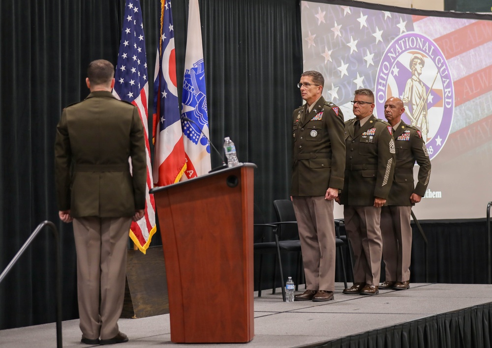 A formal military ceremony is taking place at an Ohio National Guard event, with uniformed personnel standing at attention on a stage. One officer stands at a podium area while three others are lined up on the stage in front of an Ohio National Guard backdrop. The setting appears to be an indoor venue with flags and official insignia prominently displayed.