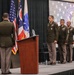 A formal military ceremony is taking place at an Ohio National Guard event, with uniformed personnel standing at attention on a stage. One officer stands at a podium area while three others are lined up on the stage in front of an Ohio National Guard backdrop. The setting appears to be an indoor venue with flags and official insignia prominently displayed.