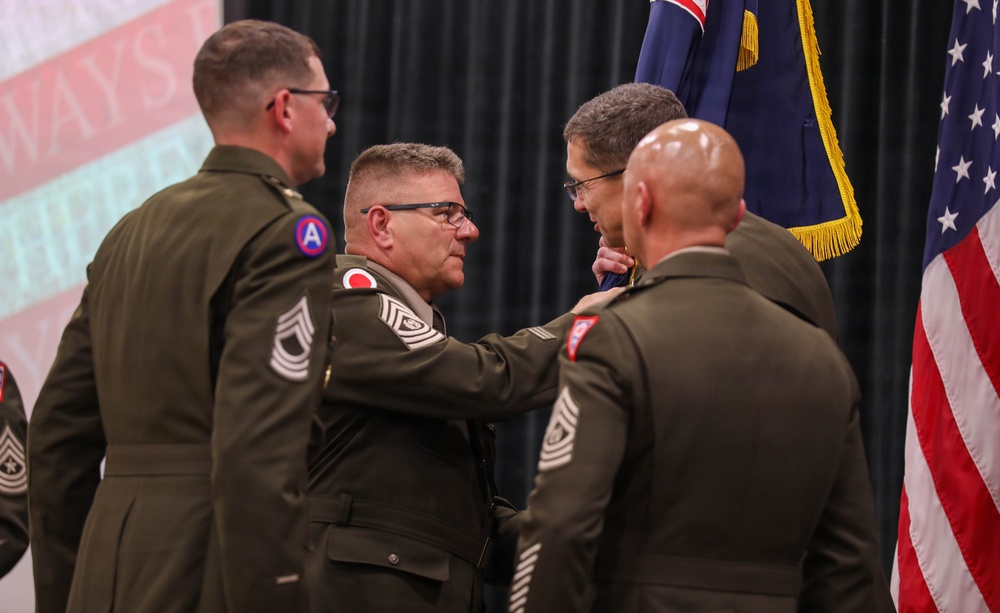 A military ceremony is taking place in which a medal or insignia is being pinned onto a soldier's uniform. Several U.S. Army personnel in olive green dress uniforms are gathered, with the central figure appearing to receive an award or decoration. An American flag and what appears to be a unit or territorial flag are visible in the background.