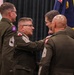 A military ceremony is taking place in which a medal or insignia is being pinned onto a soldier's uniform. Several U.S. Army personnel in olive green dress uniforms are gathered, with the central figure appearing to receive an award or decoration. An American flag and what appears to be a unit or territorial flag are visible in the background.