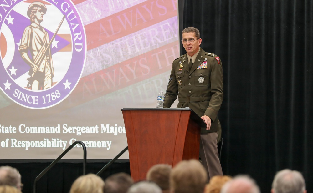 A military officer in Army Service Uniform is speaking at a podium during a formal ceremony. The event is identified as a 'State Command Sergeant Major of Responsibility Ceremony' for the Ohio National Guard, established since 1788. An audience is visible in the foreground, and a large projection screen displays the Ohio National Guard seal and the motto 'Always There.'