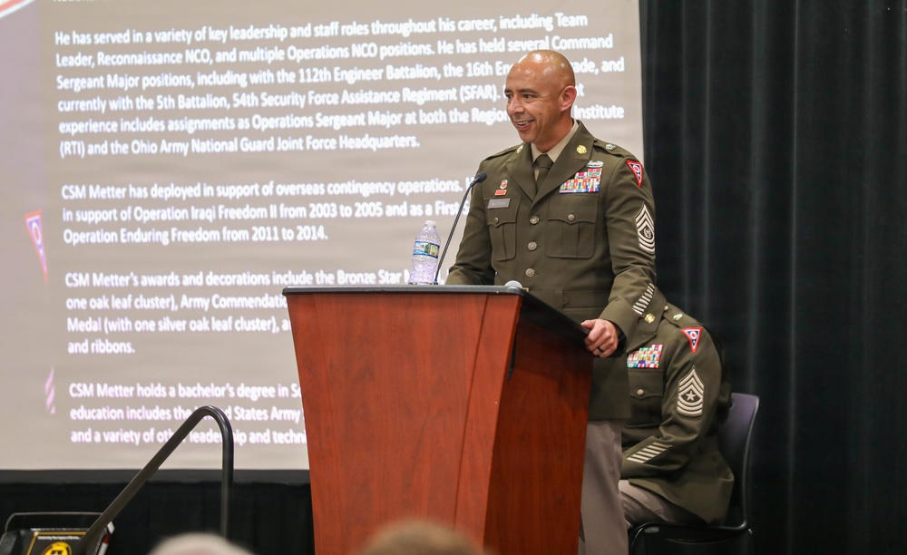 A U.S. Army Command Sergeant Major (CSM) in dress green uniform stands at a wooden podium, smiling while addressing an audience. A biographical presentation slide is projected behind him, identifying the individual as 'CSM Metter' and detailing his military career, deployments, and awards. A second uniformed soldier is partially visible seated behind the podium.