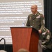 A U.S. Army Command Sergeant Major (CSM) in dress green uniform stands at a wooden podium, smiling while addressing an audience. A biographical presentation slide is projected behind him, identifying the individual as 'CSM Metter' and detailing his military career, deployments, and awards. A second uniformed soldier is partially visible seated behind the podium.