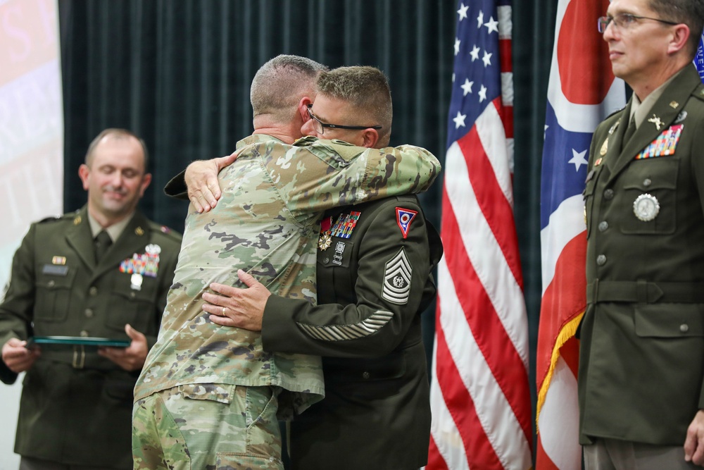 A military ceremony is taking place, likely a retirement or award ceremony, where two soldiers in U.S. Army uniforms are embracing. One soldier is wearing the Army Combat Uniform (ACU/OCP pattern) while the other is in the Army Service Uniform (ASU) with visible rank insignia of a Sergeant Major and multiple medals. Three other military personnel are present in the background, with U.S. and Ohio state flags visible.