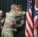 A military ceremony is taking place, likely a retirement or award ceremony, where two soldiers in U.S. Army uniforms are embracing. One soldier is wearing the Army Combat Uniform (ACU/OCP pattern) while the other is in the Army Service Uniform (ASU) with visible rank insignia of a Sergeant Major and multiple medals. Three other military personnel are present in the background, with U.S. and Ohio state flags visible.