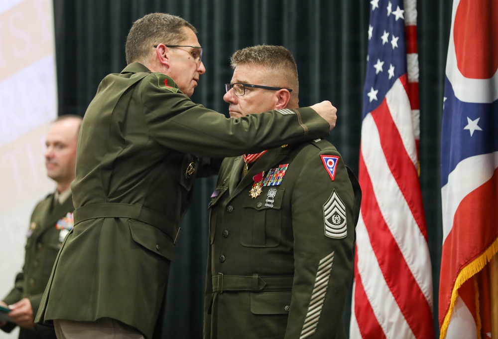 A military ceremony is taking place in which a senior officer is pinning a medal or insignia onto the collar/shoulder area of a Sergeant Major (identifiable by rank chevrons). Both individuals are wearing U.S. Army green dress uniforms, and the recipient displays multiple service ribbons and medals. The Ohio state flag and U.S. flag are visible in the background, indicating this likely involves the Ohio National Guard.