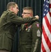 A military ceremony is taking place in which a senior officer is pinning a medal or insignia onto the collar/shoulder area of a Sergeant Major (identifiable by rank chevrons). Both individuals are wearing U.S. Army green dress uniforms, and the recipient displays multiple service ribbons and medals. The Ohio state flag and U.S. flag are visible in the background, indicating this likely involves the Ohio National Guard.
