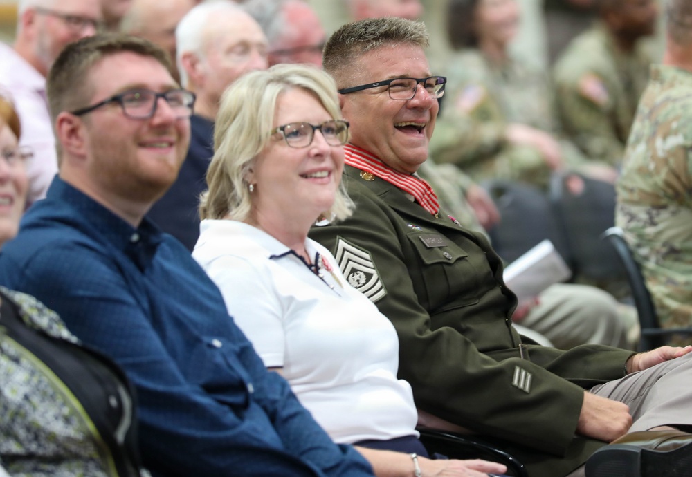 Three people seated in the foreground are laughing and smiling during what appears to be a military ceremony or event. The person on the right is wearing a military dress uniform with visible rank insignia, medals, and a red-striped collar, suggesting a senior non-commissioned officer. The background shows additional attendees, including personnel in camouflage uniforms.