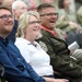 Three people seated in the foreground are laughing and smiling during what appears to be a military ceremony or event. The person on the right is wearing a military dress uniform with visible rank insignia, medals, and a red-striped collar, suggesting a senior non-commissioned officer. The background shows additional attendees, including personnel in camouflage uniforms.