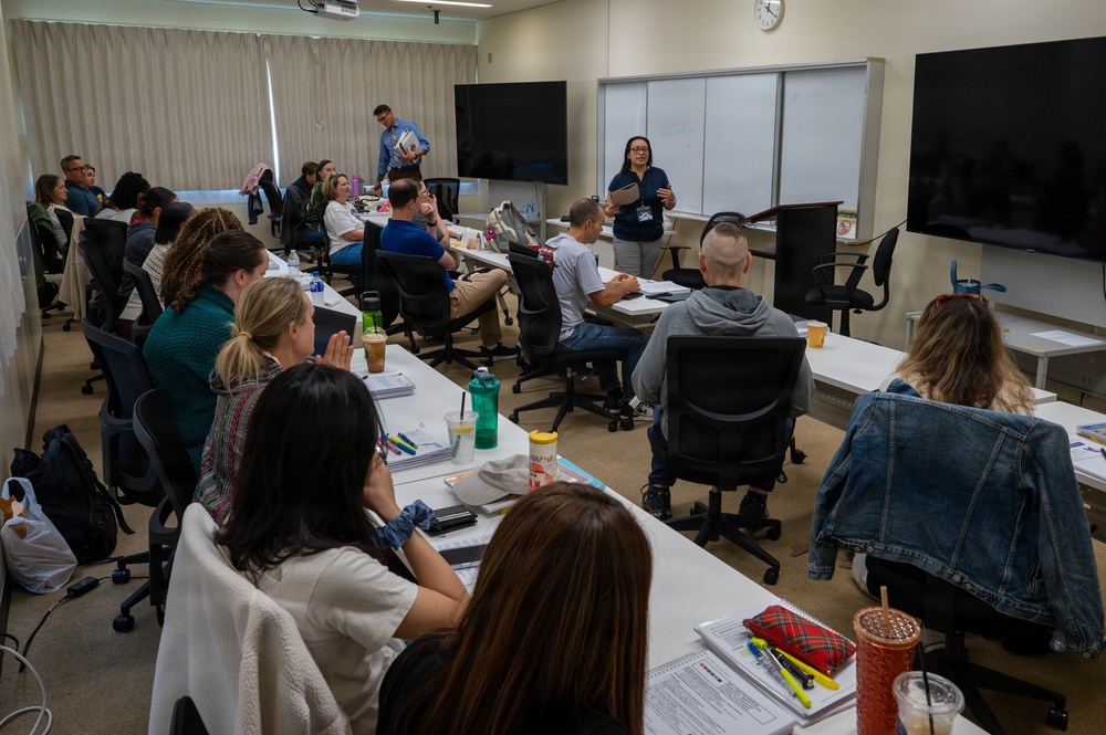 A classroom or training session is underway with approximately 20 participants seated at white tables arranged in a U-shape or rows. A female instructor wearing a navy blue shirt stands at the front near a whiteboard and large display screen, appearing to address the group. A second person in a blue shirt stands near the back of the room, also holding materials.