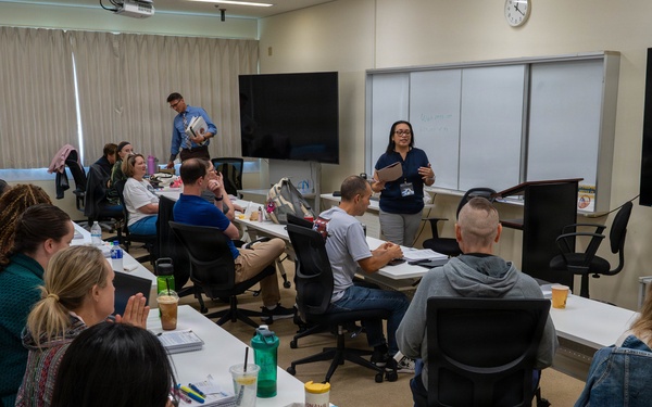 A classroom or training session is underway with approximately 20 participants seated at white tables arranged in a U-shape or rows. A female instructor wearing a navy blue shirt stands at the front near a whiteboard and large display screen, appearing to address the group. A second person in a blue shirt stands near the back of the room, also holding materials.