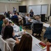 A classroom or training session is underway with approximately 20 participants seated at white tables arranged in a U-shape or rows. A female instructor wearing a navy blue shirt stands at the front near a whiteboard and large display screen, appearing to address the group. A second person in a blue shirt stands near the back of the room, also holding materials.