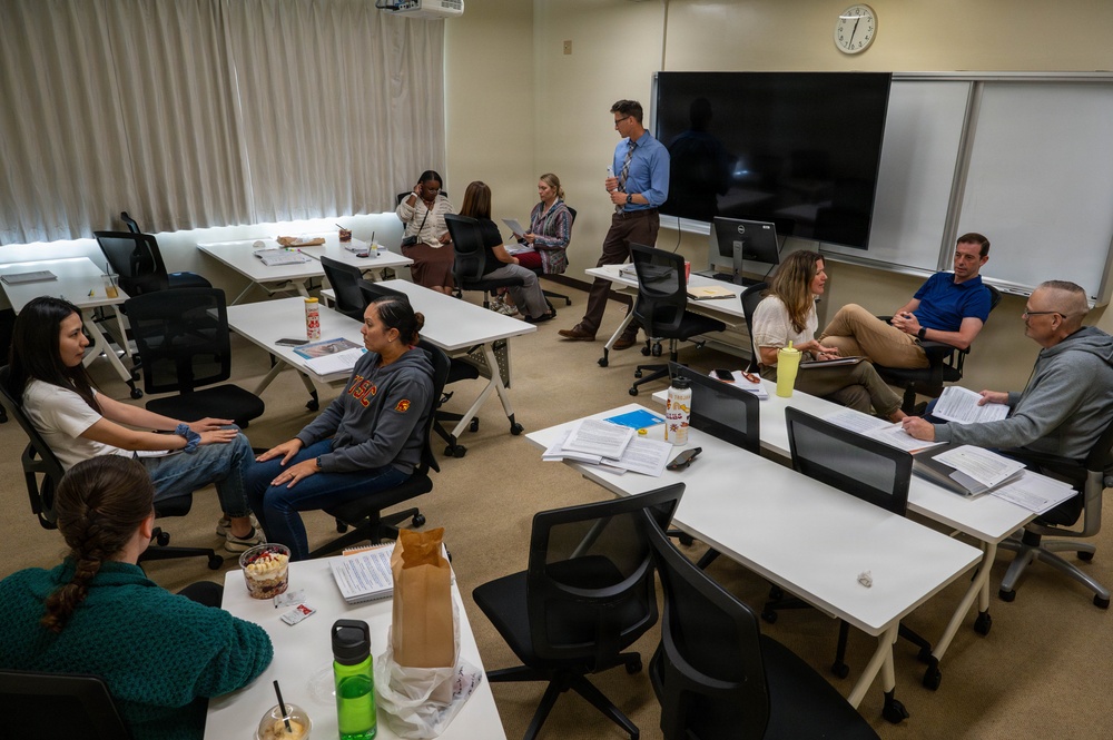 A small seminar or workshop session is taking place in a modern classroom with white modular tables and black rolling chairs. Approximately 10-11 participants are seated in a roughly circular arrangement, with one standing male presenter holding a cup near a large flat-screen display and whiteboard. The setting suggests a graduate-level class, professional training, or faculty meeting, with participants having documents, notebooks, and food/drinks on the tables.