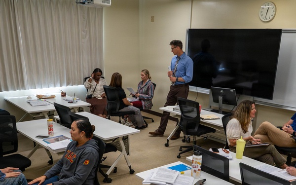 A small seminar or workshop session is taking place in a modern classroom with white modular tables and black rolling chairs. Approximately 10-11 participants are seated in a roughly circular arrangement, with one standing male presenter holding a cup near a large flat-screen display and whiteboard. The setting suggests a graduate-level class, professional training, or faculty meeting, with participants having documents, notebooks, and food/drinks on the tables.