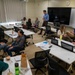 A small seminar or workshop session is taking place in a modern classroom with white modular tables and black rolling chairs. Approximately 10-11 participants are seated in a roughly circular arrangement, with one standing male presenter holding a cup near a large flat-screen display and whiteboard. The setting suggests a graduate-level class, professional training, or faculty meeting, with participants having documents, notebooks, and food/drinks on the tables.