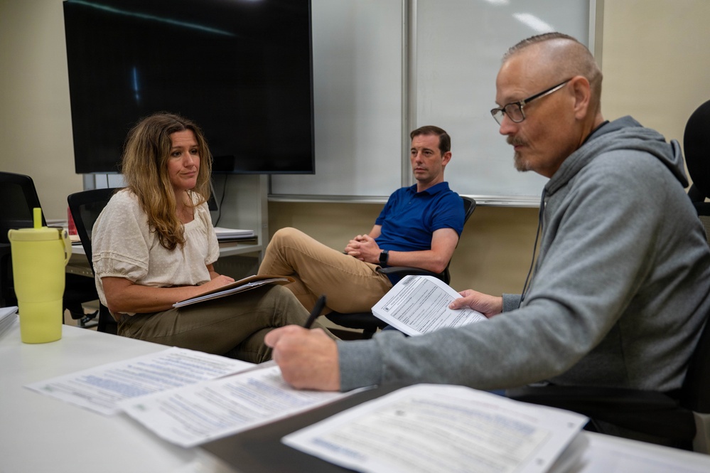 Three adults are seated in what appears to be a classroom or conference room, engaged in a discussion or meeting. A man in the foreground holds printed documents and a pen, while a woman across from him listens attentively with a folder on her lap. A second man in a blue polo shirt sits in the background observing the conversation.