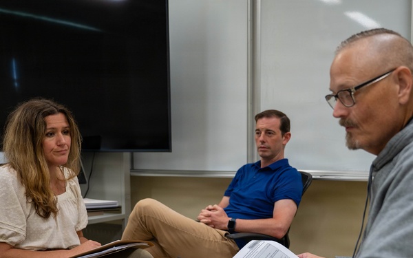 Three adults are seated in what appears to be a classroom or conference room, engaged in a discussion or meeting. A man in the foreground holds printed documents and a pen, while a woman across from him listens attentively with a folder on her lap. A second man in a blue polo shirt sits in the background observing the conversation.