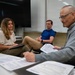 Three adults are seated in what appears to be a classroom or conference room, engaged in a discussion or meeting. A man in the foreground holds printed documents and a pen, while a woman across from him listens attentively with a folder on her lap. A second man in a blue polo shirt sits in the background observing the conversation.