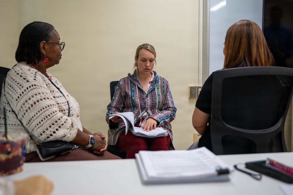 Three women are seated in what appears to be a professional meeting or consultation room. The central figure is reviewing documents in a binder or folder, while the other two women appear to be listening or observing. The setting suggests a formal discussion, interview, or case review session.
