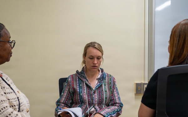 Three women are seated in what appears to be a professional meeting or consultation room. The central figure is reviewing documents in a binder or folder, while the other two women appear to be listening or observing. The setting suggests a formal discussion, interview, or case review session.