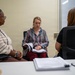 Three women are seated in what appears to be a professional meeting or consultation room. The central figure is reviewing documents in a binder or folder, while the other two women appear to be listening or observing. The setting suggests a formal discussion, interview, or case review session.