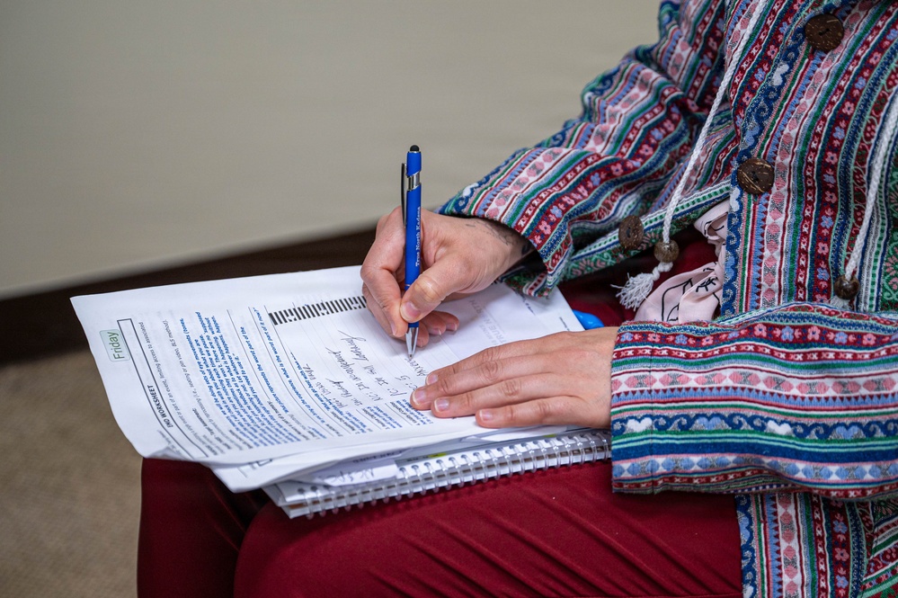 A person wearing a colorful, patterned ethnic-style jacket is writing on a printed worksheet or form placed on top of a spiral notebook. The document appears to be labeled 'Friday' and contains structured text fields. The person is using a blue pen and appears to be filling out the form while seated.