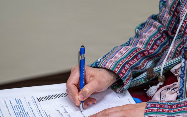 A person wearing a colorful, patterned ethnic-style jacket is writing on a printed worksheet or form placed on top of a spiral notebook. The document appears to be labeled 'Friday' and contains structured text fields. The person is using a blue pen and appears to be filling out the form while seated.