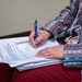 A person wearing a colorful, patterned ethnic-style jacket is writing on a printed worksheet or form placed on top of a spiral notebook. The document appears to be labeled 'Friday' and contains structured text fields. The person is using a blue pen and appears to be filling out the form while seated.