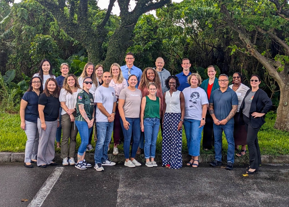A large group of approximately 25 adults poses together outdoors in what appears to be a tropical or subtropical setting, likely for an organizational or professional group photo. The group is diverse in age, gender, and ethnicity, standing in two rows in a parking lot area. Lush green trees and vegetation form the backdrop behind them.