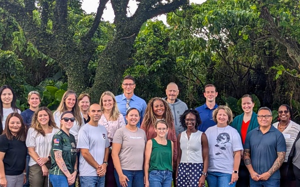 A large group of approximately 25 adults poses together outdoors in what appears to be a tropical or subtropical setting, likely for an organizational or professional group photo. The group is diverse in age, gender, and ethnicity, standing in two rows in a parking lot area. Lush green trees and vegetation form the backdrop behind them.