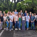 A large group of approximately 25 adults poses together outdoors in what appears to be a tropical or subtropical setting, likely for an organizational or professional group photo. The group is diverse in age, gender, and ethnicity, standing in two rows in a parking lot area. Lush green trees and vegetation form the backdrop behind them.