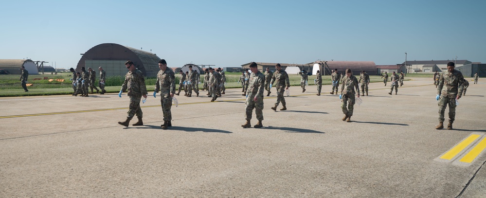 A large group of military personnel in camouflage uniforms are conducting a foreign object debris (FOD) walk on an airfield tarmac. They are spread out in a line formation, walking slowly and looking downward to collect debris from the runway surface. Several aircraft hangars and base structures are visible in the background.