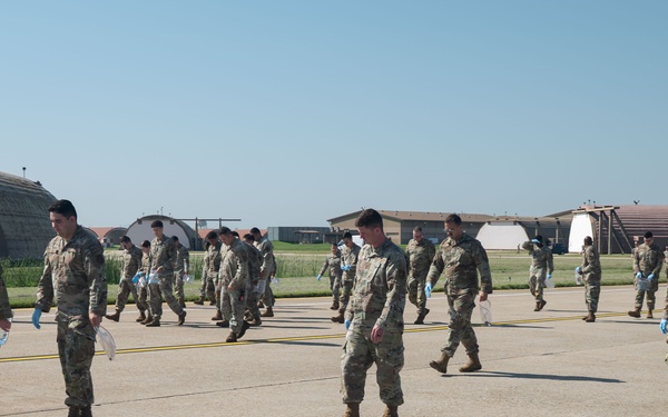 A large group of military personnel in camouflage uniforms are conducting a foreign object debris (FOD) walk on an airfield tarmac. They are spread out in a line formation, walking slowly and looking downward to collect debris from the runway surface. Several aircraft hangars and base structures are visible in the background.