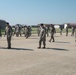 A large group of military personnel in camouflage uniforms are conducting a foreign object debris (FOD) walk on an airfield tarmac. They are spread out in a line formation, walking slowly and looking downward to collect debris from the runway surface. Several aircraft hangars and base structures are visible in the background.