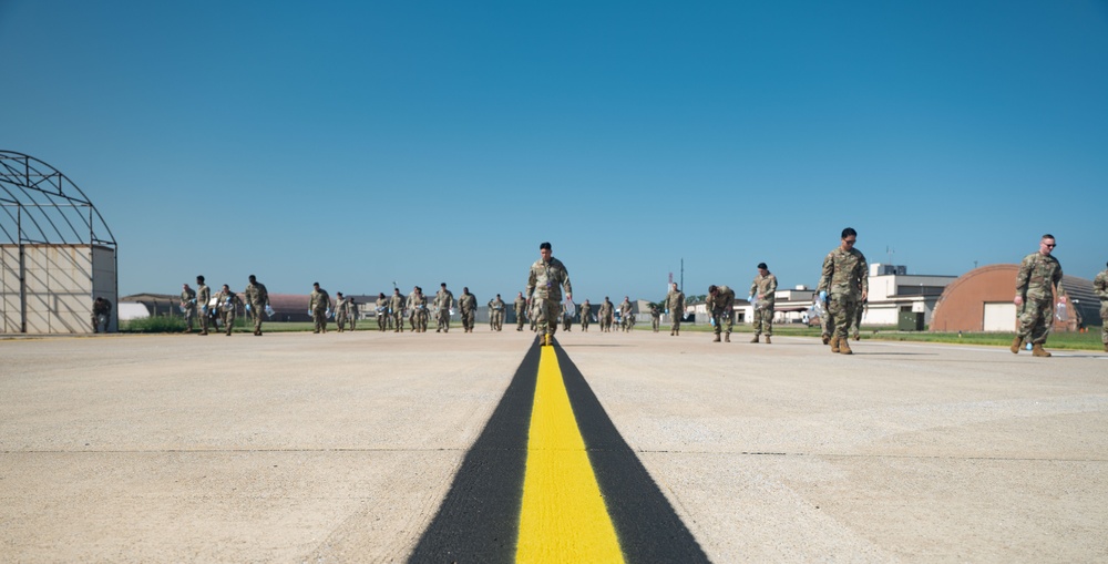 A large group of military personnel in camouflage uniforms are walking across an airfield tarmac toward the camera. The scene is set at a military air base with hangars visible in the background under a clear blue sky. A prominent yellow centerline stripe runs along the concrete surface in the foreground.