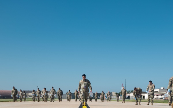 A large group of military personnel in camouflage uniforms are walking across an airfield tarmac toward the camera. The scene is set at a military air base with hangars visible in the background under a clear blue sky. A prominent yellow centerline stripe runs along the concrete surface in the foreground.