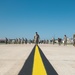 A large group of military personnel in camouflage uniforms are walking across an airfield tarmac toward the camera. The scene is set at a military air base with hangars visible in the background under a clear blue sky. A prominent yellow centerline stripe runs along the concrete surface in the foreground.
