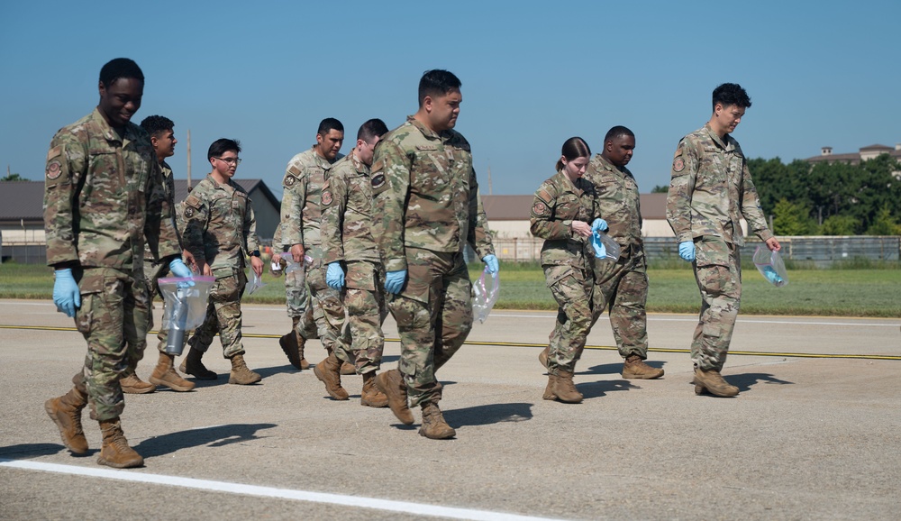 A group of military personnel in OCP (Operational Camouflage Pattern) uniforms are walking across what appears to be a flight line or tarmac at a military installation. Several individuals are wearing blue nitrile gloves and carrying clear plastic bags, suggesting they are conducting a Foreign Object Debris (FOD) walk, a common airfield safety procedure. The setting appears to be a U.S. Air Force base based on uniform insignia and airfield infrastructure.