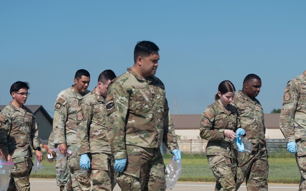 A group of military personnel in OCP (Operational Camouflage Pattern) uniforms are walking across what appears to be a flight line or tarmac at a military installation. Several individuals are wearing blue nitrile gloves and carrying clear plastic bags, suggesting they are conducting a Foreign Object Debris (FOD) walk, a common airfield safety procedure. The setting appears to be a U.S. Air Force base based on uniform insignia and airfield infrastructure.