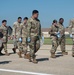 A group of military personnel in OCP (Operational Camouflage Pattern) uniforms are walking across what appears to be a flight line or tarmac at a military installation. Several individuals are wearing blue nitrile gloves and carrying clear plastic bags, suggesting they are conducting a Foreign Object Debris (FOD) walk, a common airfield safety procedure. The setting appears to be a U.S. Air Force base based on uniform insignia and airfield infrastructure.
