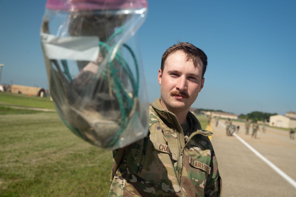 A U.S. Air Force servicemember, identified by the name tape beginning with 'CHA' and 'U.S. AIR FORCE' tape on their OCP (Operational Camouflage Pattern) uniform, holds up a clear plastic bag containing unidentified items toward the camera. The photo is taken outdoors on what appears to be a military installation, with a flight line or road, grass, and buildings visible in the background. Other personnel in uniform can be seen in the background.