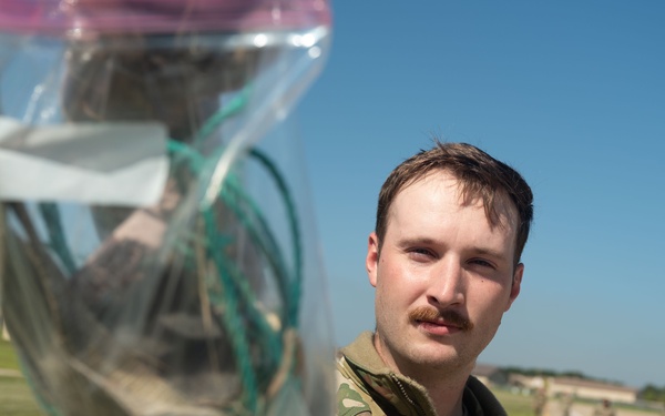 A U.S. Air Force servicemember, identified by the name tape beginning with 'CHA' and 'U.S. AIR FORCE' tape on their OCP (Operational Camouflage Pattern) uniform, holds up a clear plastic bag containing unidentified items toward the camera. The photo is taken outdoors on what appears to be a military installation, with a flight line or road, grass, and buildings visible in the background. Other personnel in uniform can be seen in the background.