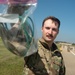 A U.S. Air Force servicemember, identified by the name tape beginning with 'CHA' and 'U.S. AIR FORCE' tape on their OCP (Operational Camouflage Pattern) uniform, holds up a clear plastic bag containing unidentified items toward the camera. The photo is taken outdoors on what appears to be a military installation, with a flight line or road, grass, and buildings visible in the background. Other personnel in uniform can be seen in the background.
