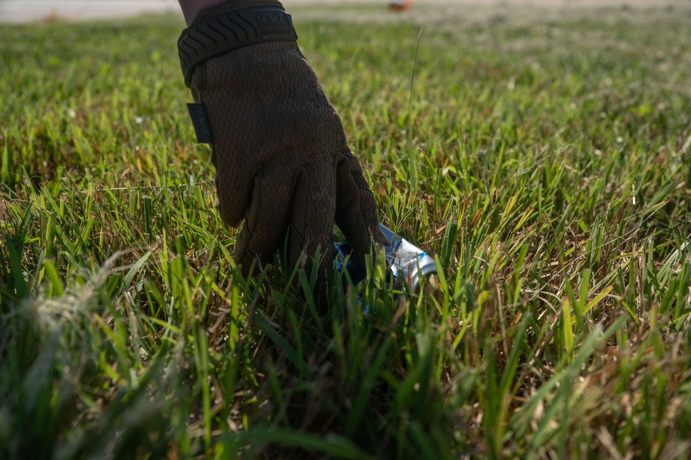 A gloved hand is reaching down to pick up a piece of litter from a grassy area. The glove is dark brown/black and appears to be a tactical or work-style glove. This appears to document a cleanup or litter removal activity.