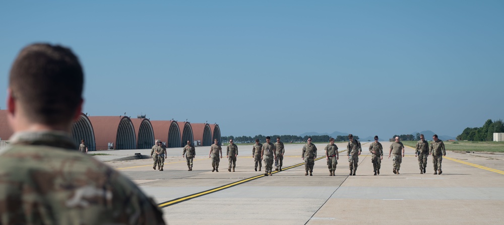 A group of approximately 12-15 military personnel in camouflage uniforms are walking in a line across an airfield tarmac, observed by a soldier in the foreground whose back is to the camera. The setting appears to be a military air base featuring distinctive arched aircraft shelters or hardened aircraft shelters (HAS) in the background. The scene suggests a formation walk, foreign object debris (FOD) walk, or post-mission assembly.