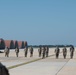 A group of approximately 12-15 military personnel in camouflage uniforms are walking in a line across an airfield tarmac, observed by a soldier in the foreground whose back is to the camera. The setting appears to be a military air base featuring distinctive arched aircraft shelters or hardened aircraft shelters (HAS) in the background. The scene suggests a formation walk, foreign object debris (FOD) walk, or post-mission assembly.