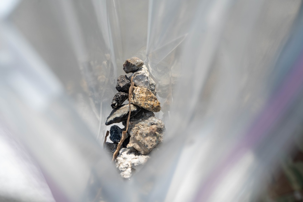 The image shows a collection of small rocks and stones of varying colors and textures, visible through a transparent plastic bag or sheet. The rocks range from dark black/charcoal to light gray and tan/yellow hues, with what appears to be a small dried twig or plant stem among them. The plastic material creates a soft, diffused framing effect around the central subject.