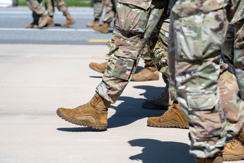 The image shows a close-up view of military personnel marching in formation on a paved surface. Multiple soldiers are visible from approximately the waist down, wearing matching camouflage uniforms and tan combat boots. The tight framing emphasizes the uniformity and coordinated movement of the group.